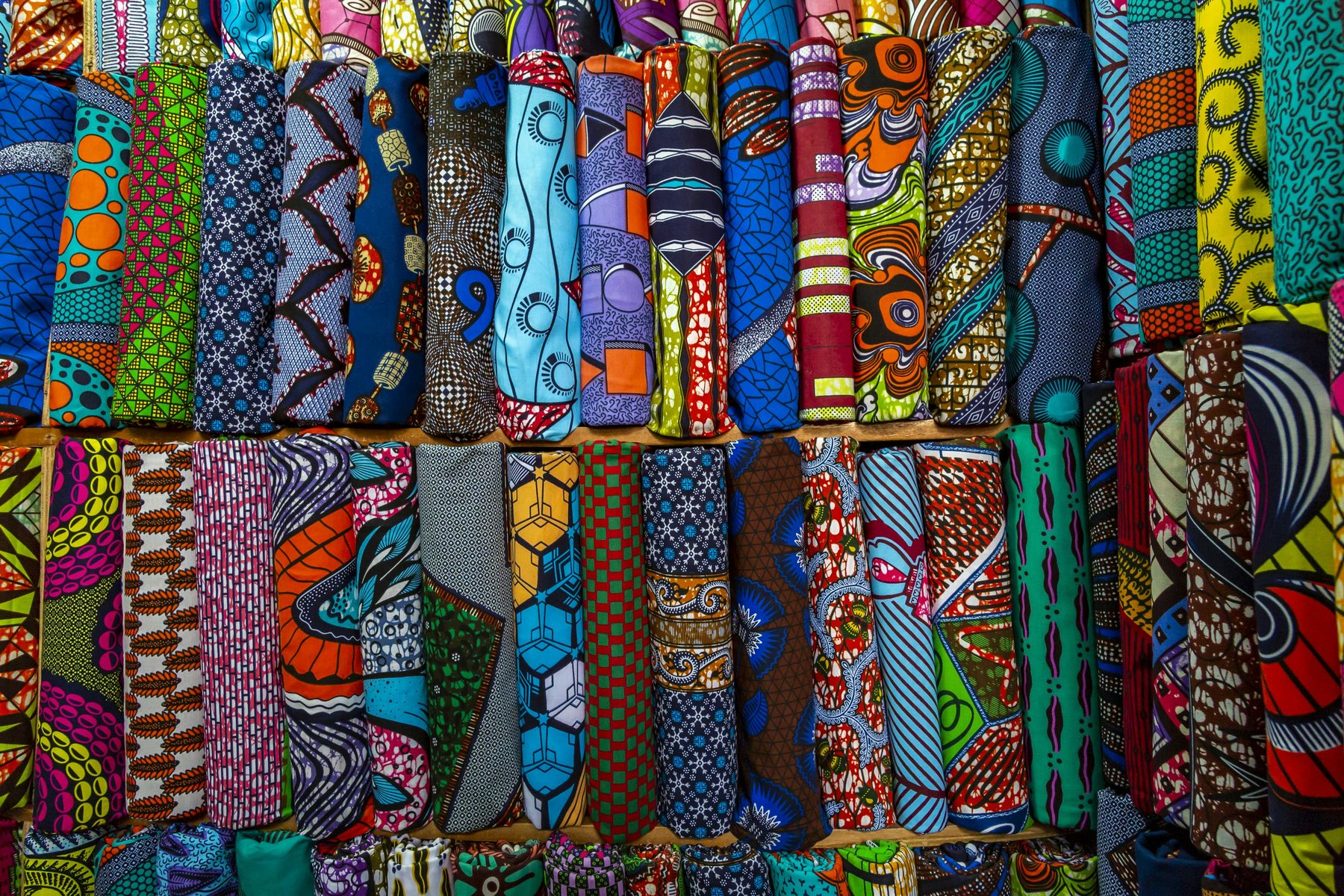 Rolls of colourful wax-print cotton fabric in bold patterns stacked on wooden market shelves