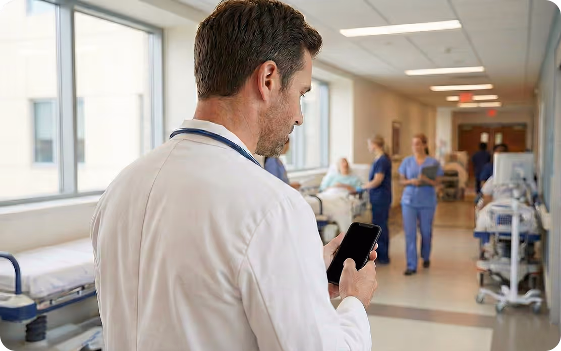 Un médecin portant une blouse blanche consulte un smartphone dans un couloir d'hôpital avec du personnel médical et des patients en arrière-plan.