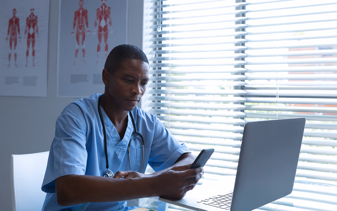 Médecin en blouse bleue assis devant un ordinateur portable, regardant son téléphone dans un bureau avec des posters anatomiques en arrière-plan.