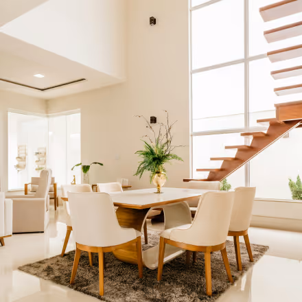 Bright, modern dining area with wooden table, six white cushioned chairs, a vase with greenery centerpiece, and floating wooden stairs beside large windows.