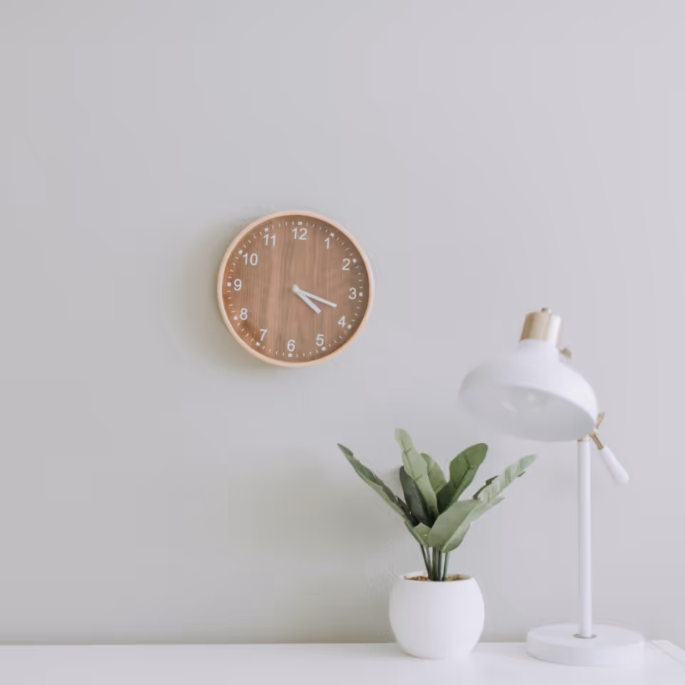 Wooden wall clock showing 3:19 above a white desk with a green potted plant and a white desk lamp.