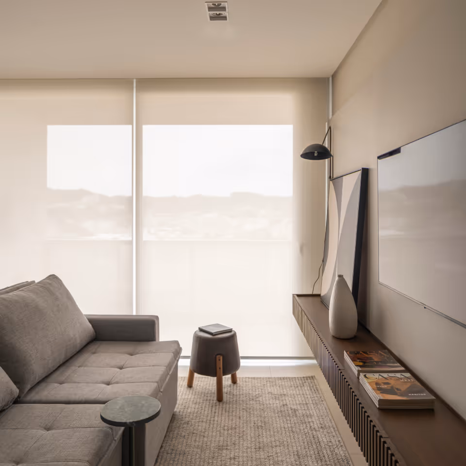 Modern living room with gray sofa, small round table, stool, wall-mounted TV, and minimalistic decor on a long shelf.