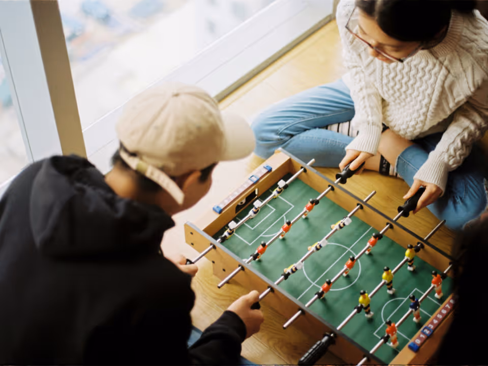 Two people playing foosball on a table near a window, one wearing a beige cap and black hoodie, the other in a white sweater and blue jeans.