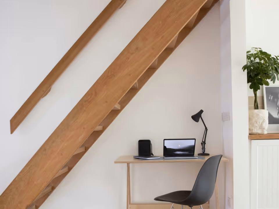 Minimalist wooden desk with laptop, notebook, lamp, and black chair under angled wooden stairs in a bright room.