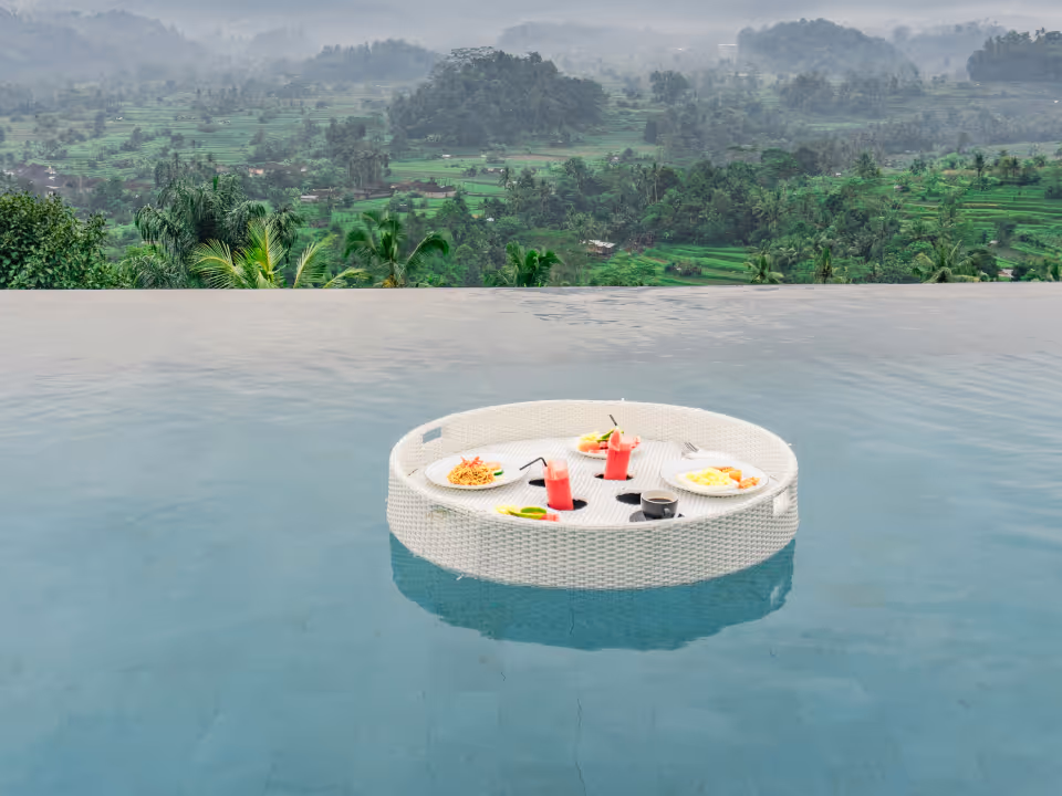 Floating round tray with plates of food and pink drinks in an infinity pool overlooking a lush green valley.