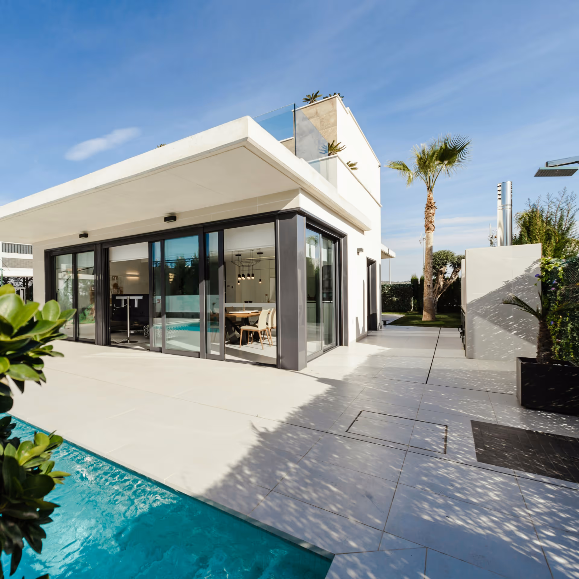 Modern white house with large glass sliding doors opening to a patio and swimming pool, with palm trees and clear sky.