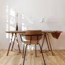 Minimalist wooden dining table with a brown chair, wine bottle, and folded cloth against a white wall with window light.