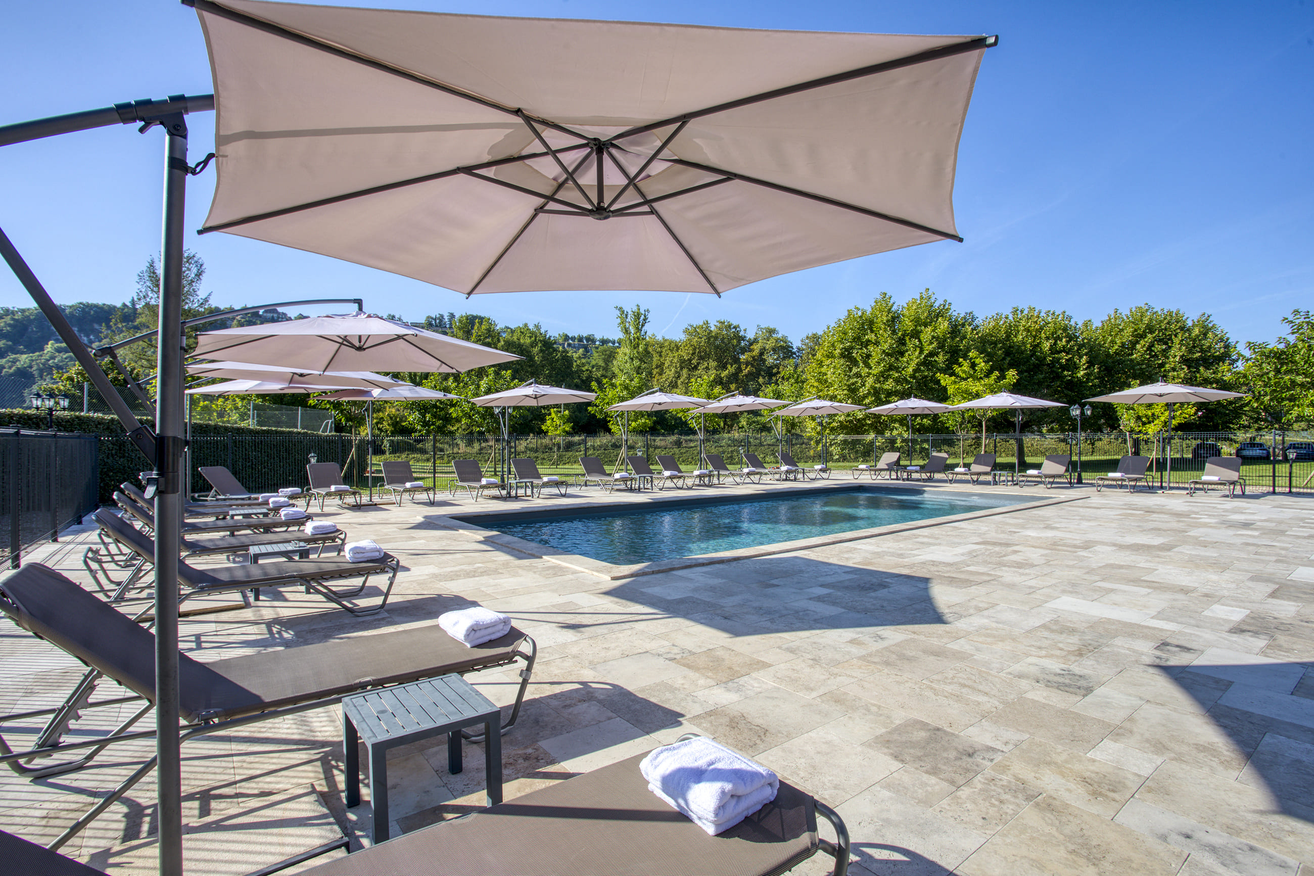 Piscine extérieure entourée de chaises longues avec serviettes blanches et grands parasols sous un ciel bleu.