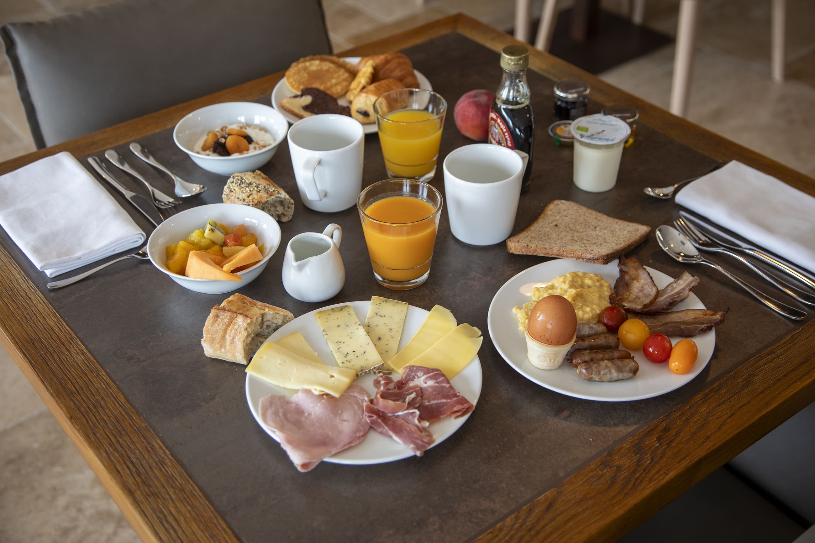 Petit déjeuner complet avec assiettes de charcuterie, fromages, œufs brouillés, saucisses, bacon, fruits, pain, yaourt, jus d'orange et café sur une table en bois.