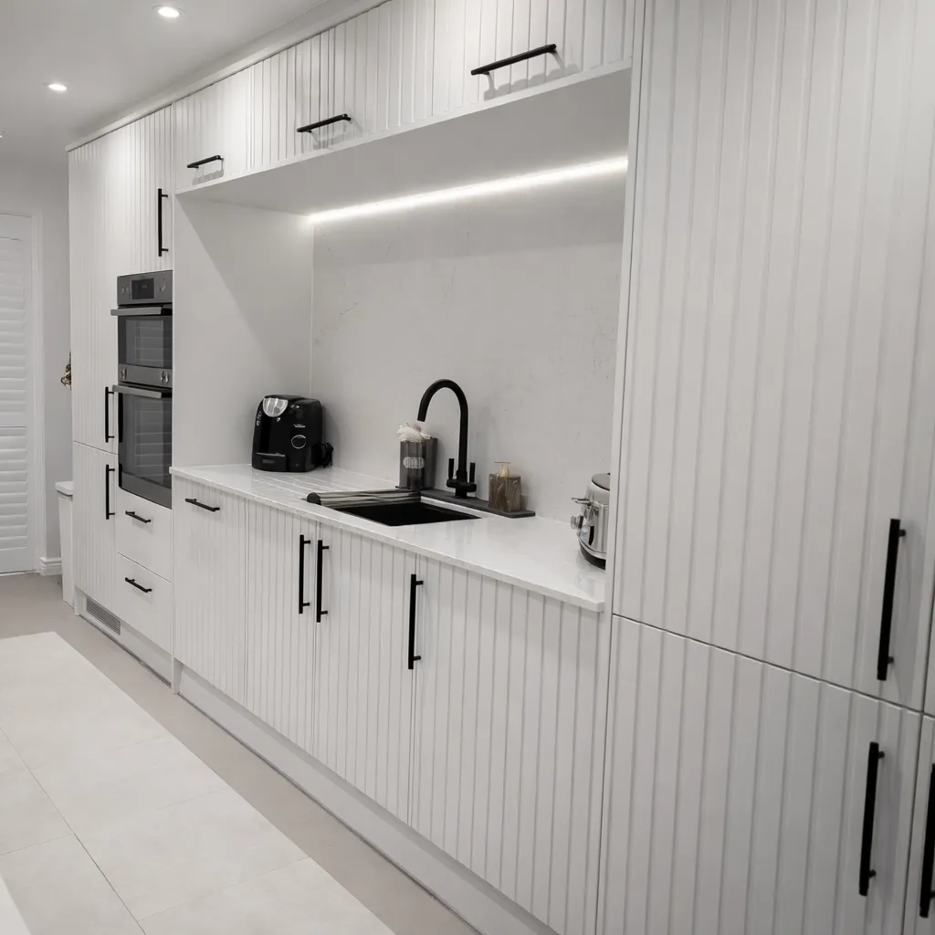 Modern white kitchen with vertical panel cabinets, black handles, under-cabinet lighting, black faucet, and countertop appliances.