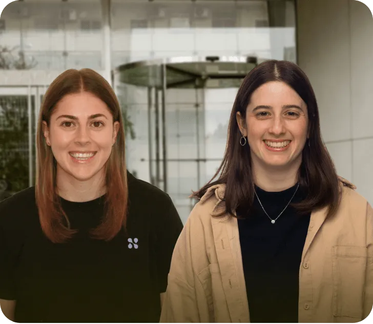 Two smiling women with straight hair standing indoors in front of a glass revolving door.