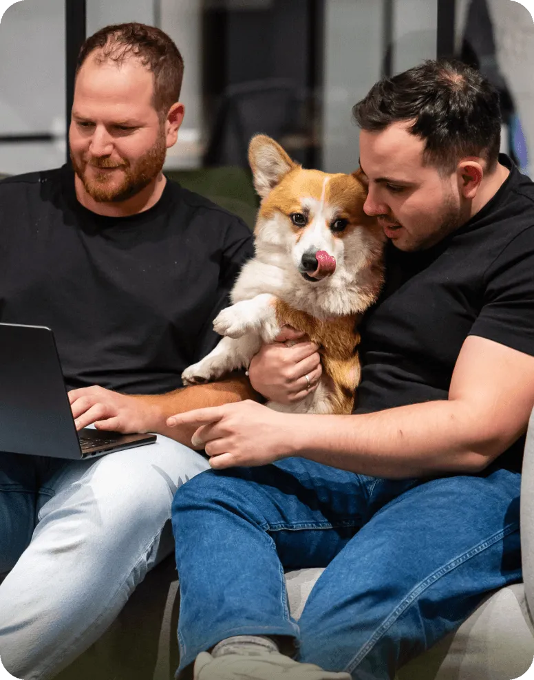 Two men sitting on a couch, one holding a corgi dog while the other points at a laptop screen.