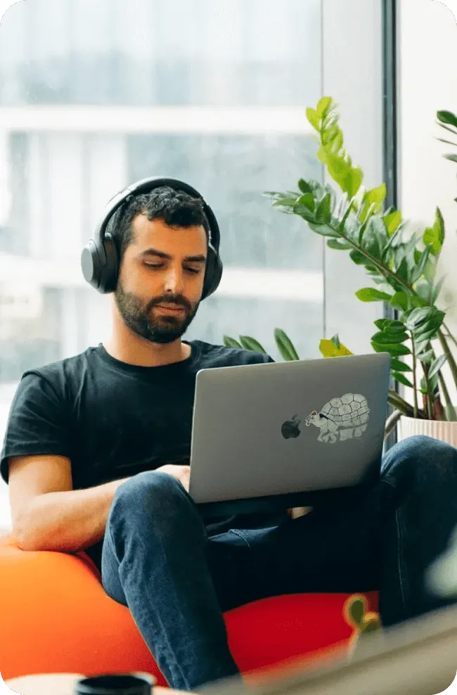 Man wearing headphones sitting on an orange bean bag working on a laptop with a turtle sticker near a window with green plants.