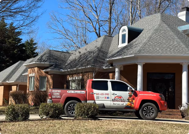 Red and white pickup truck with Brothers Roofing ads parked on driveway of a brick house with a grey roof.