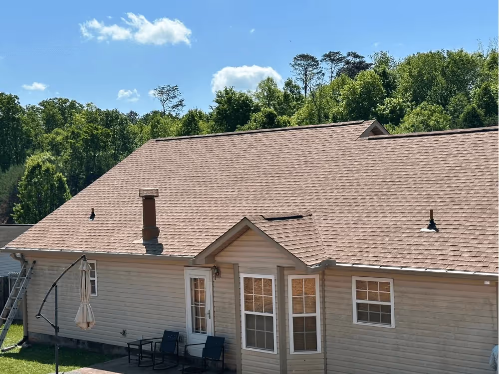Single-story house with a beige shingle roof, beige siding, patio door, and two black patio chairs under a blue sky with clouds and green trees in the background.