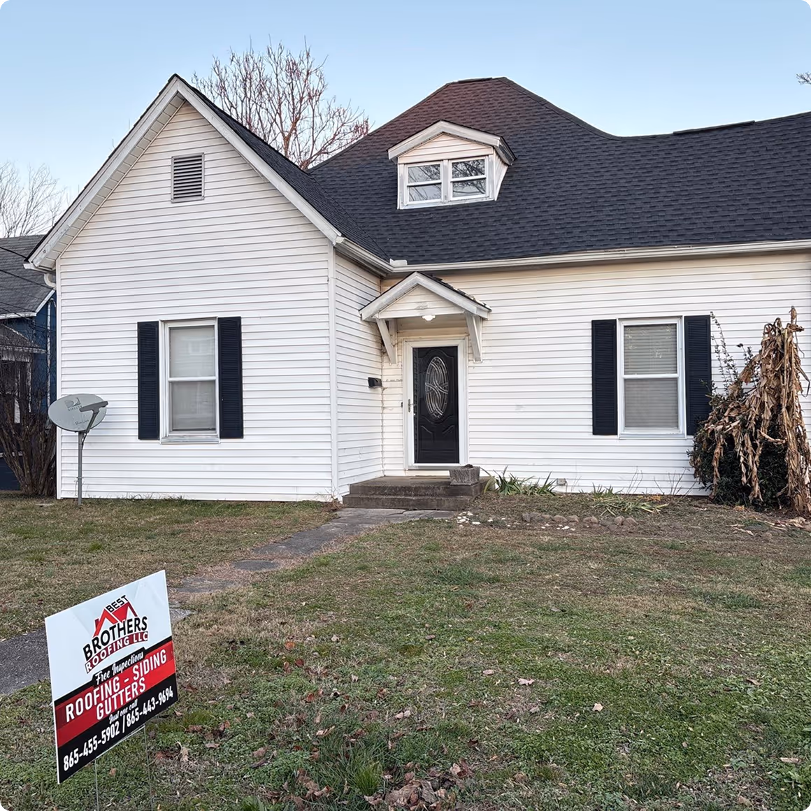 White house with black shutters, a black front door with an ornate oval glass panel, and a gabled dormer window on a black roof, with a lawn sign advertising roofing, siding, and gutters services by Best Brothers Roofing LLC.
