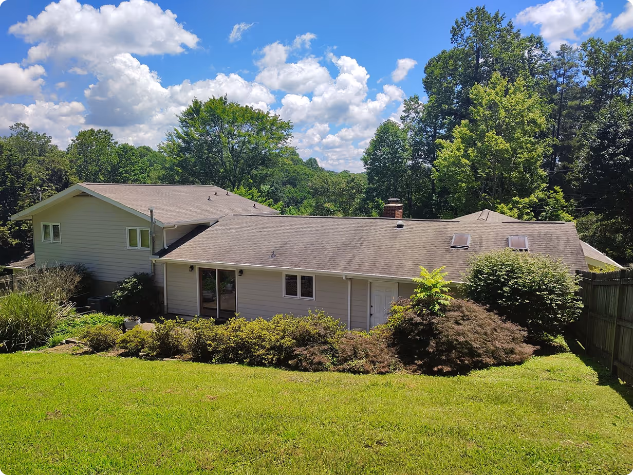 Single-story beige house with brown roof behind a green lawn and shrubs under a blue sky with scattered clouds.