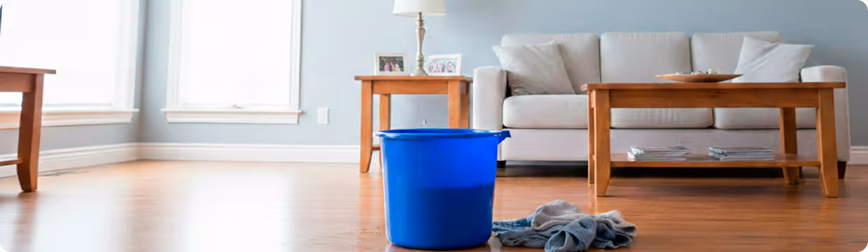 Blue bucket collecting water from a leak on a wooden floor in a living room with a gray sofa and wooden tables.