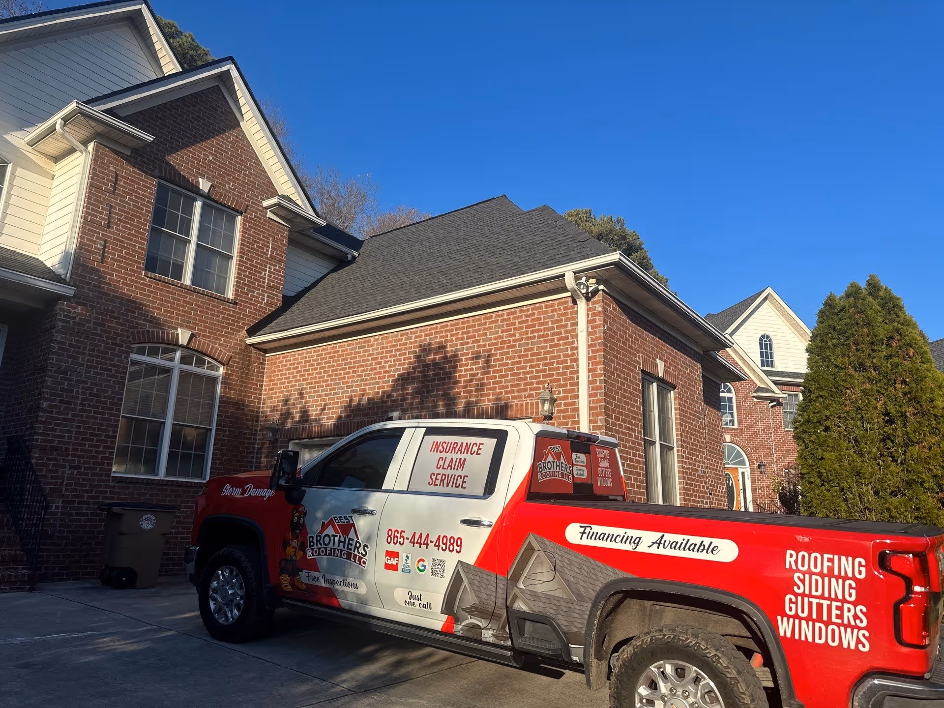 Red and white Best Brothers Roofing LLC service truck parked in driveway beside a brick house, advertising roofing, siding, gutters, windows, and insurance claim service.
