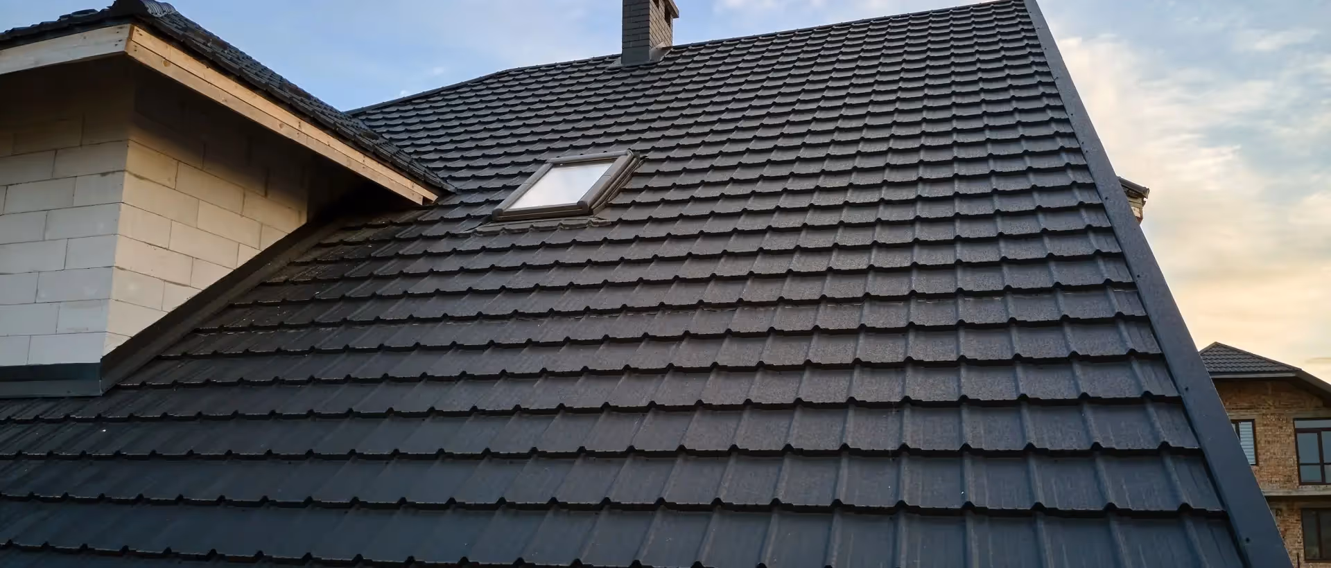 Black tiled roof with a small skylight window and a chimney under a partly cloudy sky.