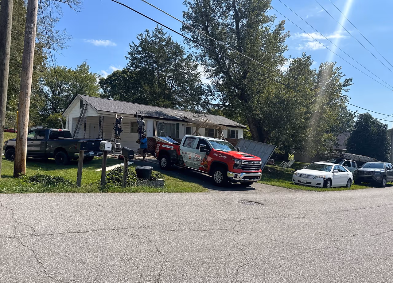 Workers repairing the exterior of a white single-story house with ladders and a red and white service truck parked in the driveway.