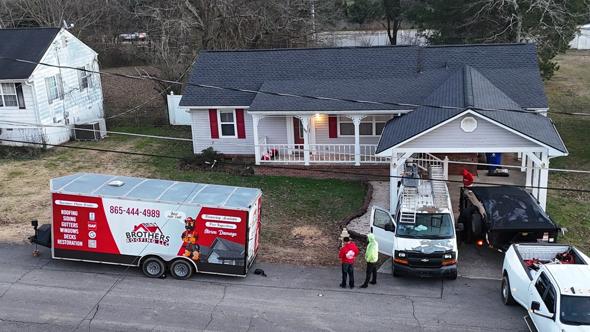 Small house with a porch, two workers near a white van and a trailer with roofing company branding.