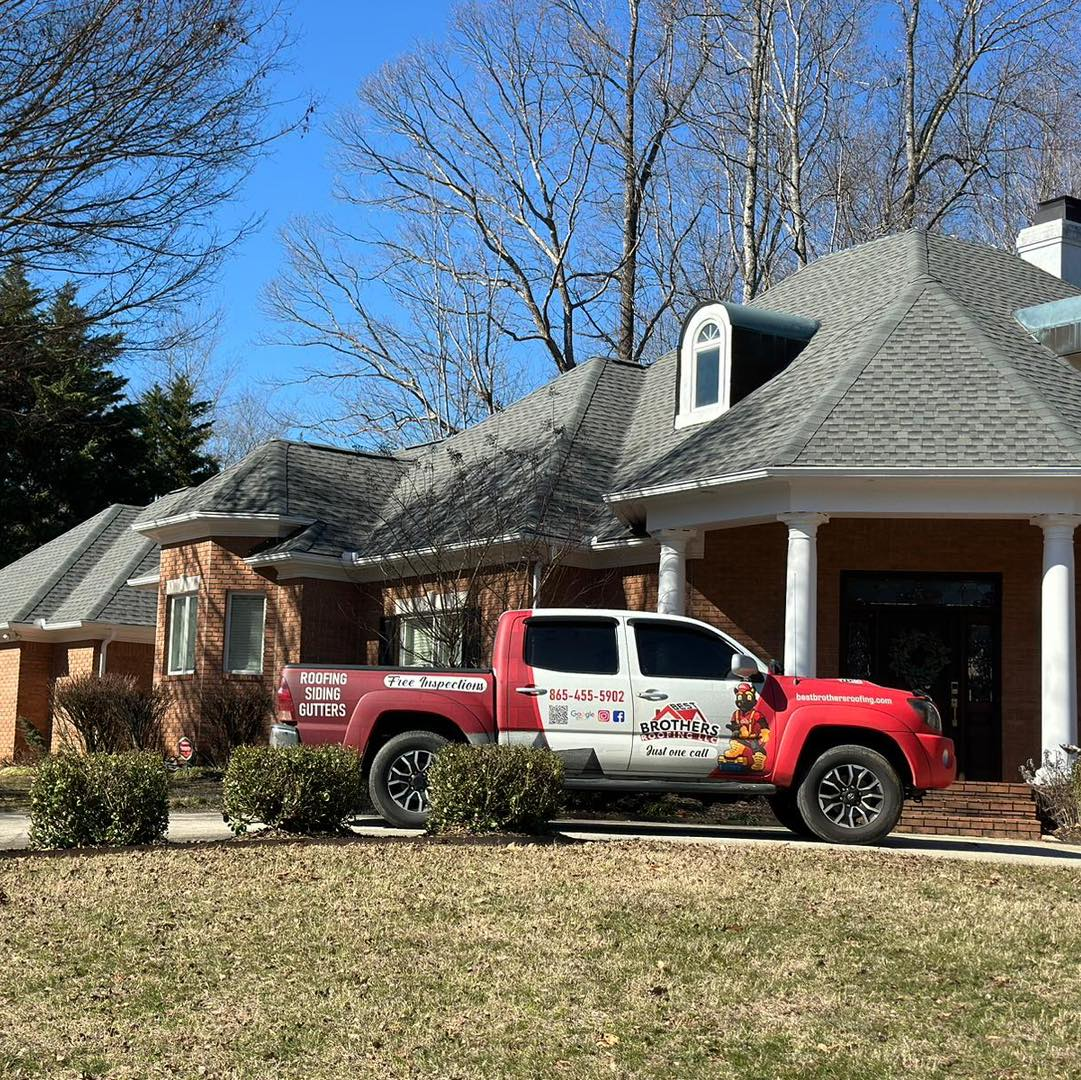 Red and white Brothers Roofing truck parked in front of a brick house with gray shingle roof.