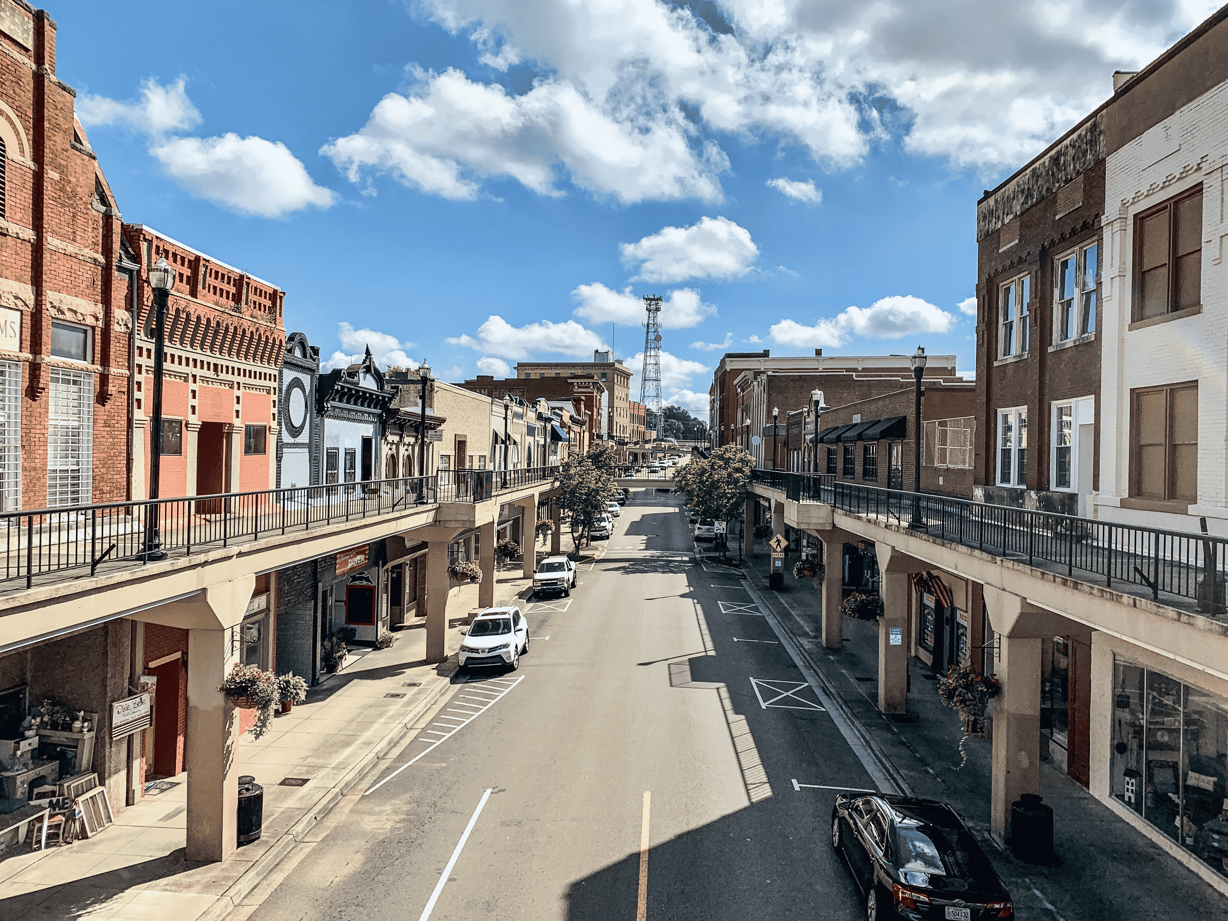 Downtown street view with historic brick buildings on both sides, elevated walkways, parked cars, trees, and a blue sky with scattered clouds.