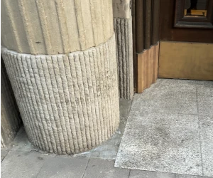 Close-up of a fluted stone column base next to a textured stone floor and wooden wall corner.
