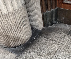 Close-up of textured stone columns and tiled ground near a wooden door corner.