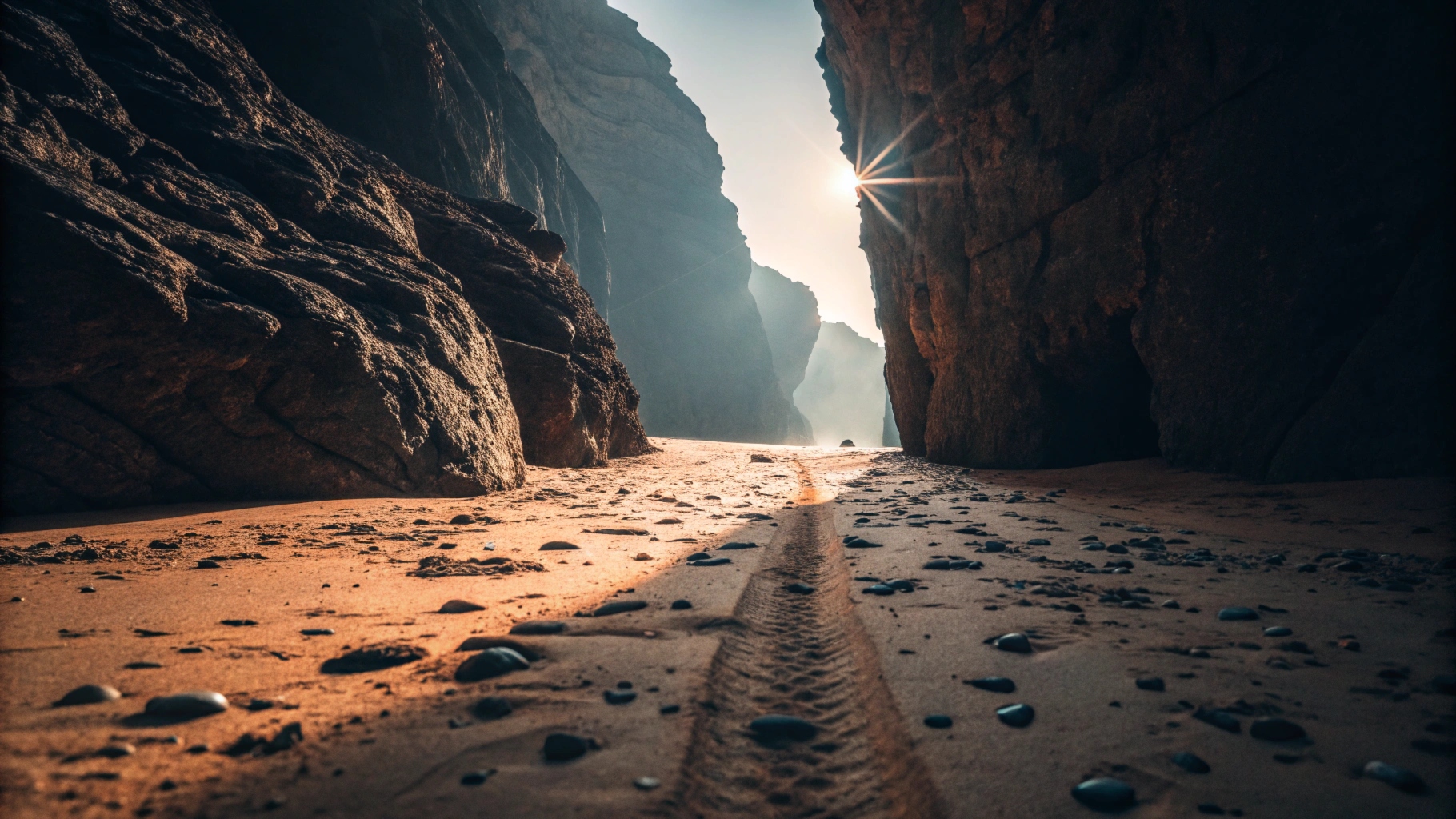 Sonnenlicht fällt durch einen schmalen Spalt zwischen hohen Felsen auf einen sandigen Boden mit Steinen und einer Reifenspur, die in die Ferne führt.