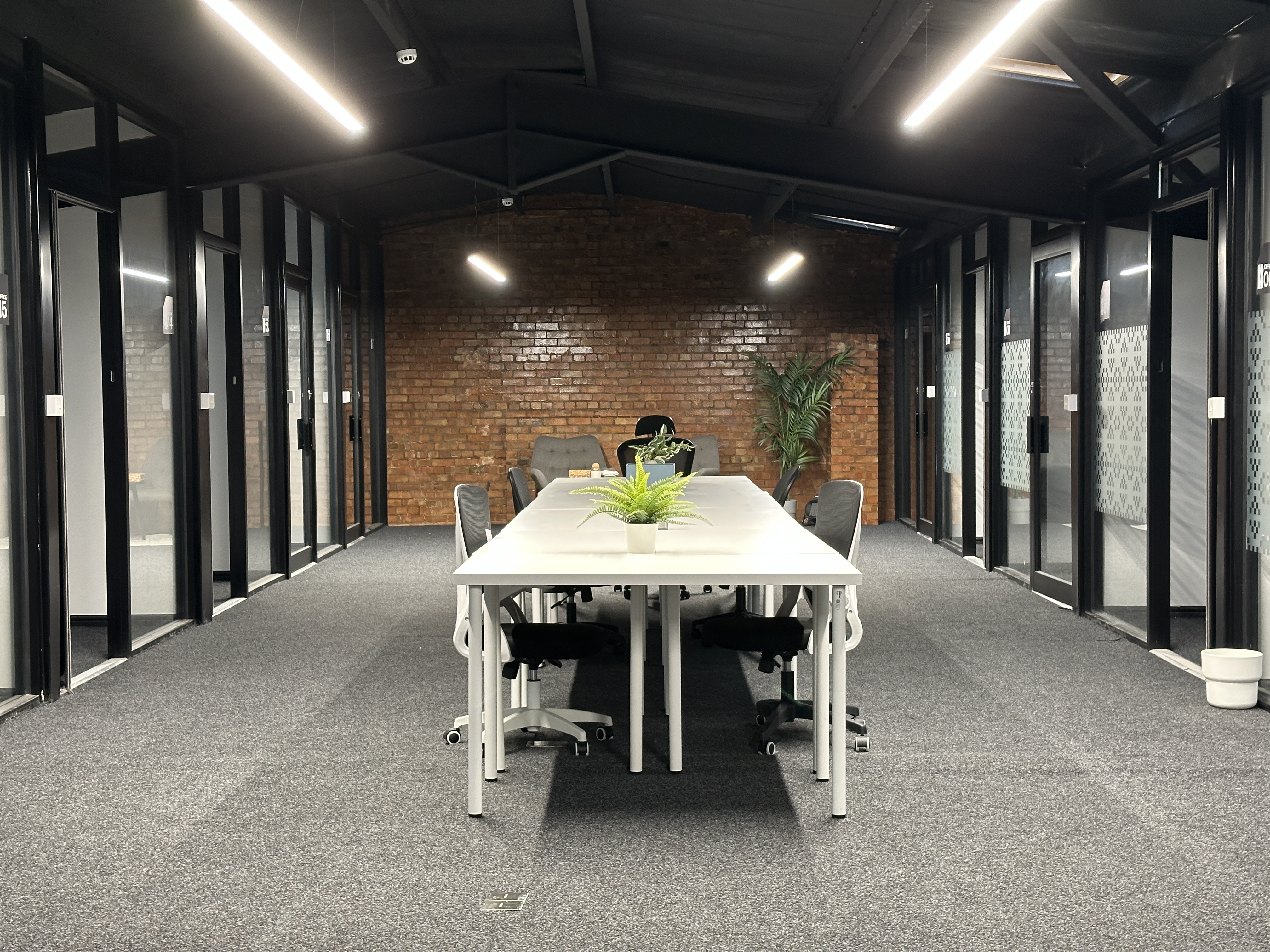 Modern office space with white desks arranged in two rows, black chairs, potted plants, and a brick wall background.