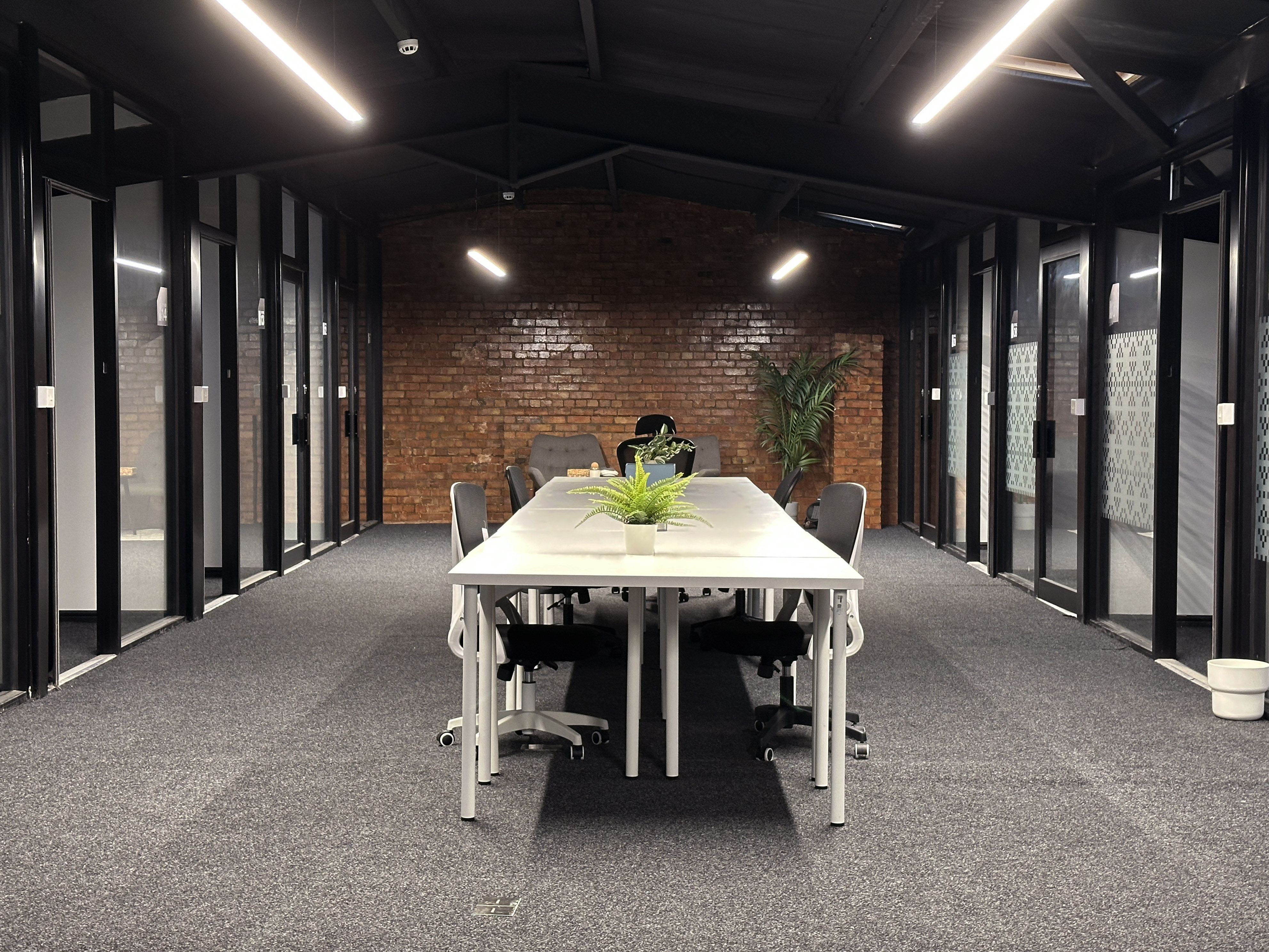 Modern office meeting room with a long white table, black office chairs, potted plants, glass-walled rooms, and exposed brick wall.