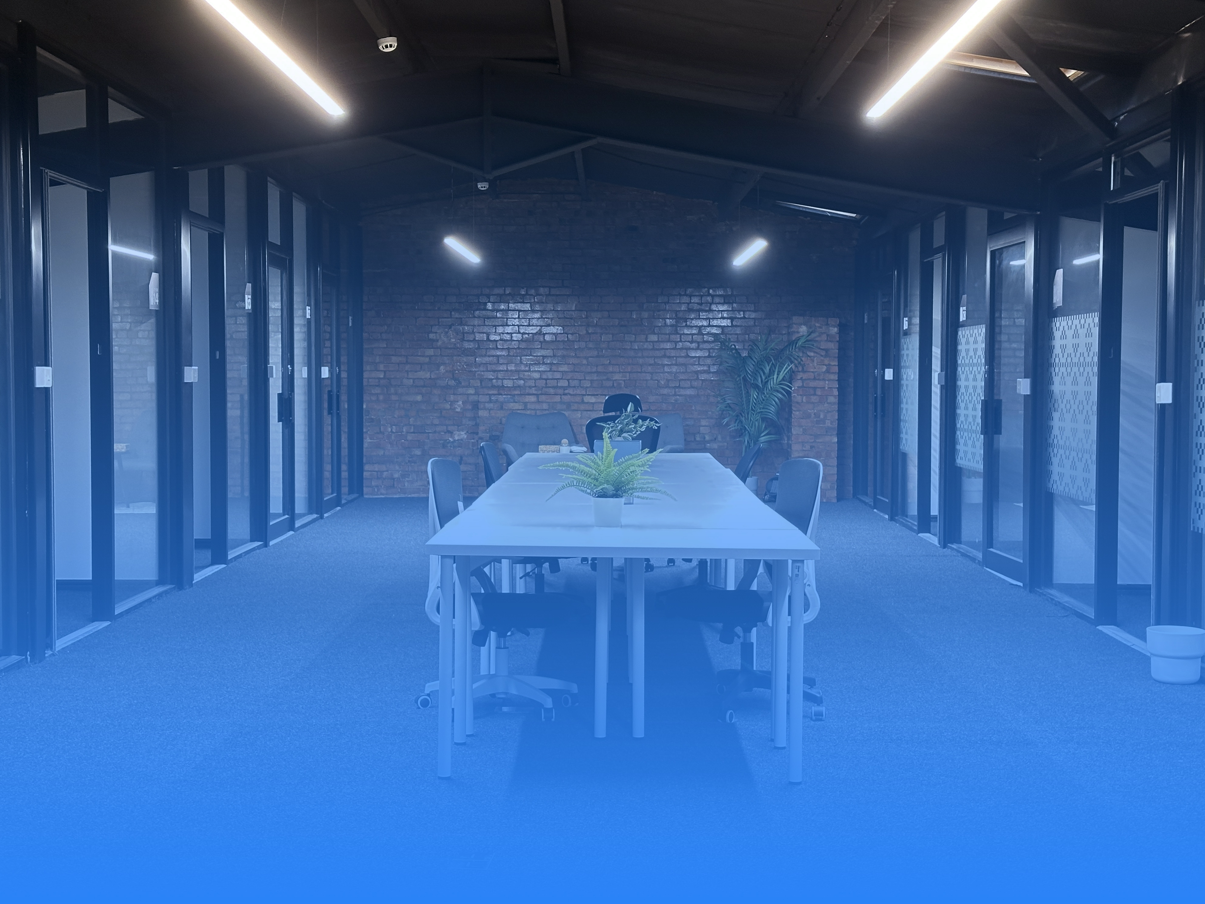 Modern office meeting room with a long white table, several gray chairs, potted plants, exposed brick wall, and glass-walled offices along the sides.
