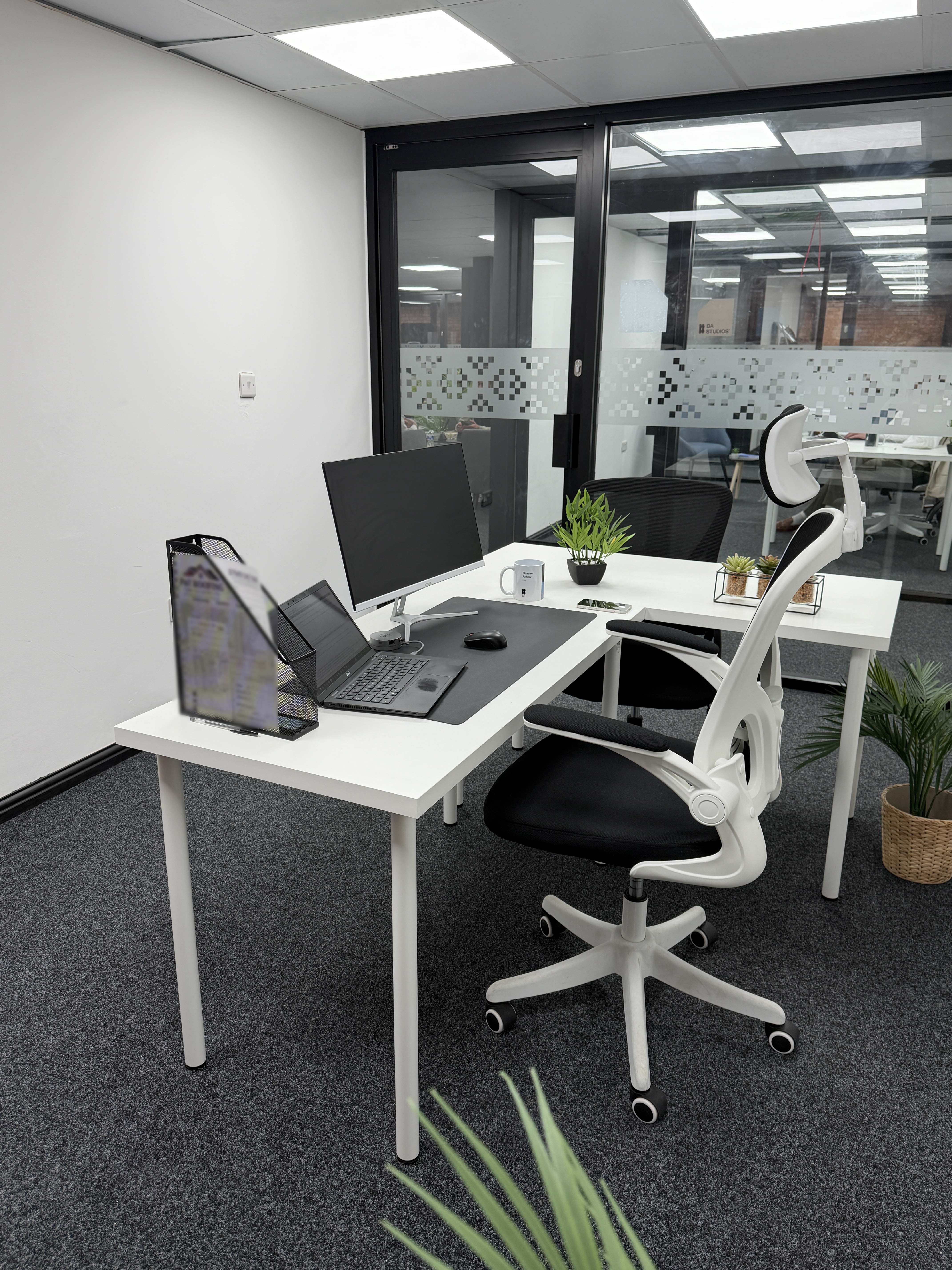 Modern office desk with a laptop, monitor, mouse, potted plants, and ergonomic chair on gray carpet near glass wall.