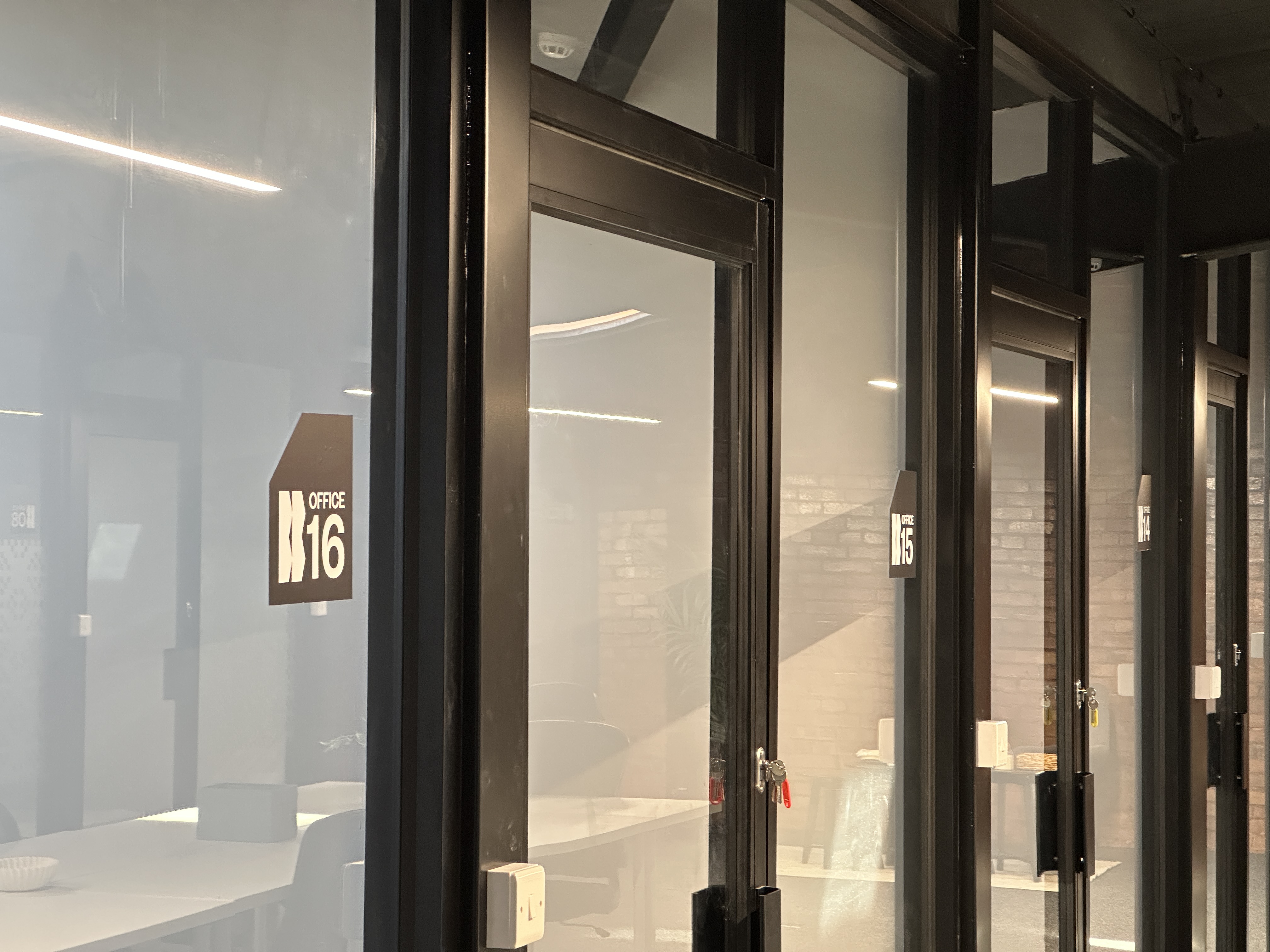 Glass office doors in a modern workspace labeled Office 14, Office 15, and Office 16 with black frames and keys in locks.