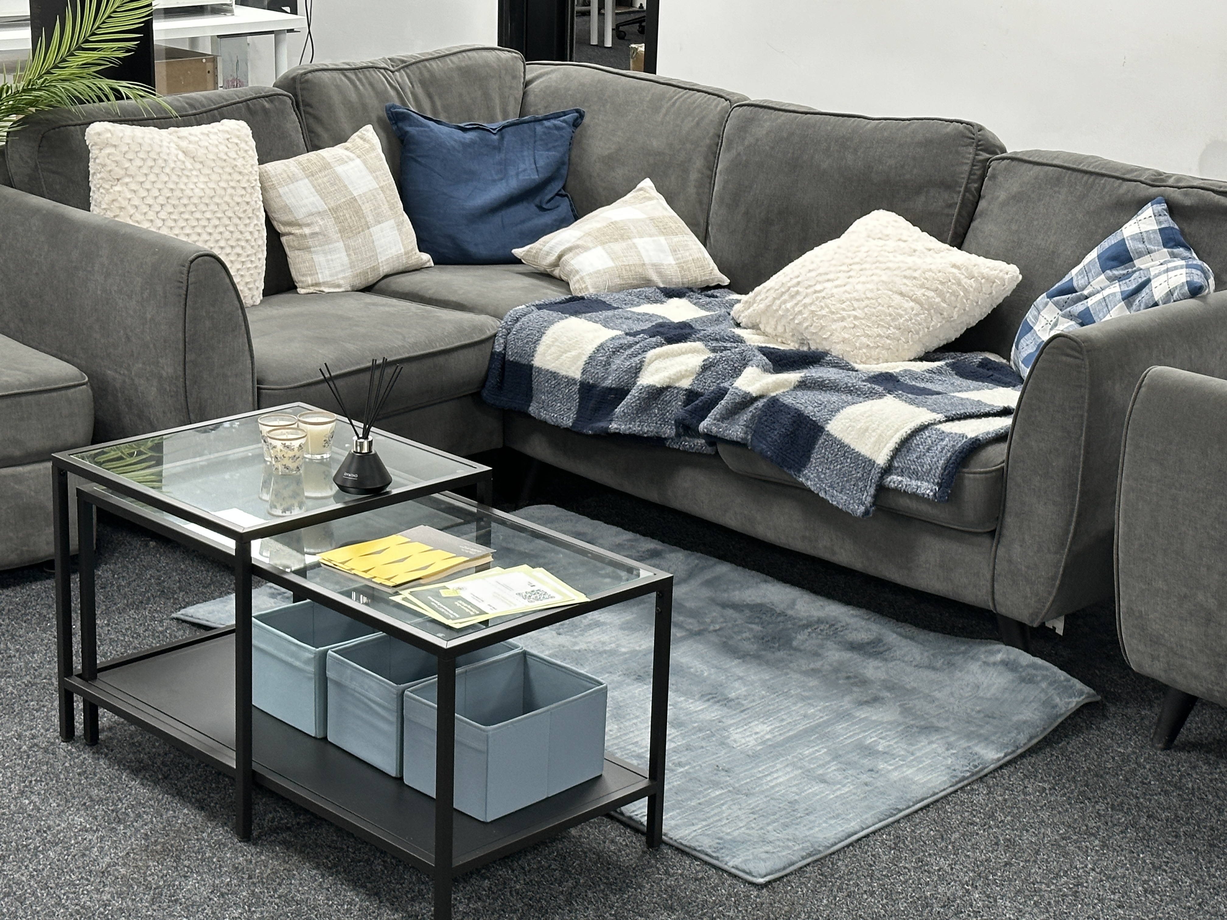Gray sectional sofa with blue and cream pillows and a blue checkered throw blanket, next to a glass coffee table with candles, reed diffuser, and storage boxes underneath.