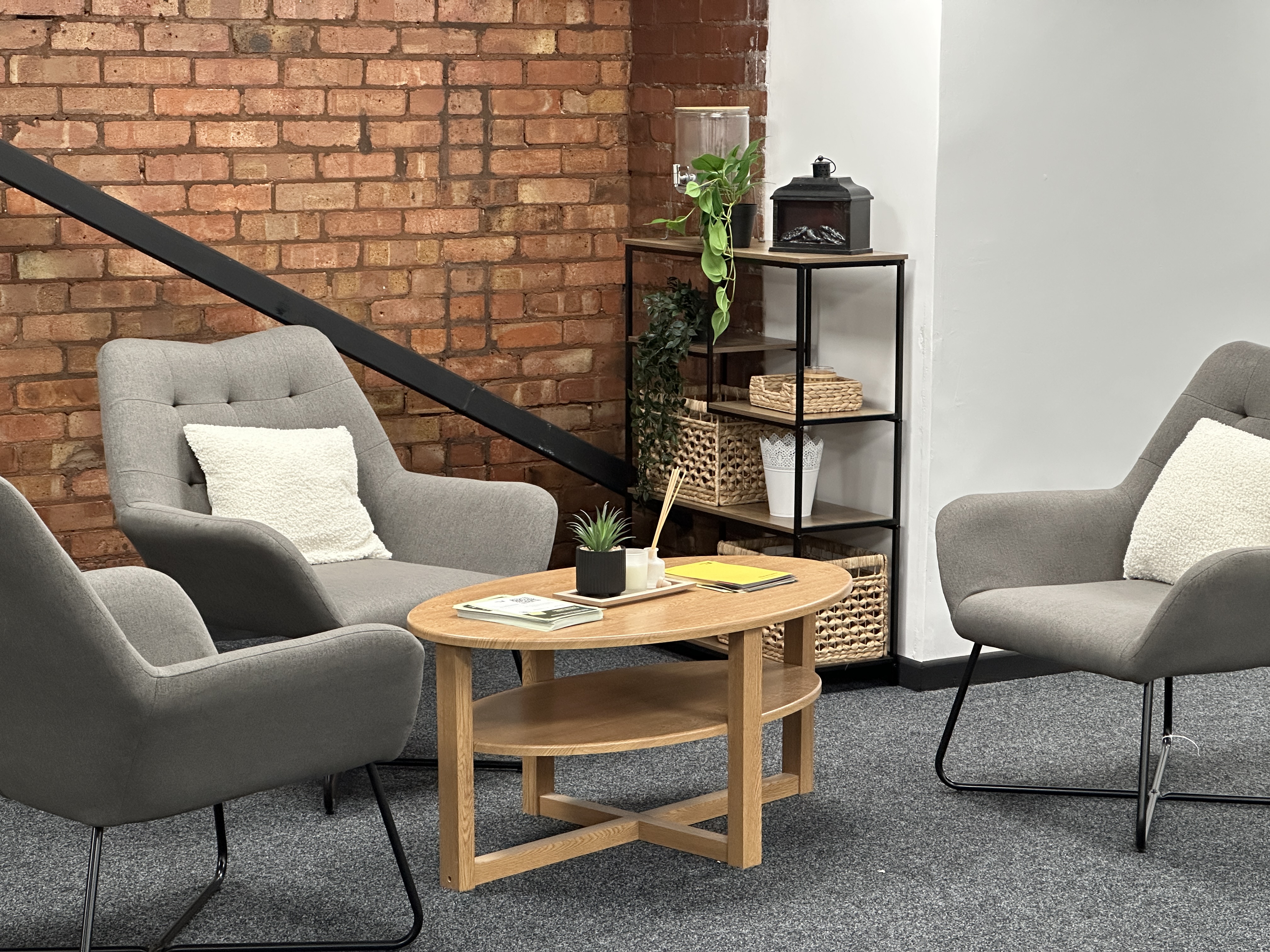 Modern sitting area with two gray armchairs featuring white cushions, a wooden coffee table, and a black metal shelving unit with plants and wicker baskets against a brick and white walls.