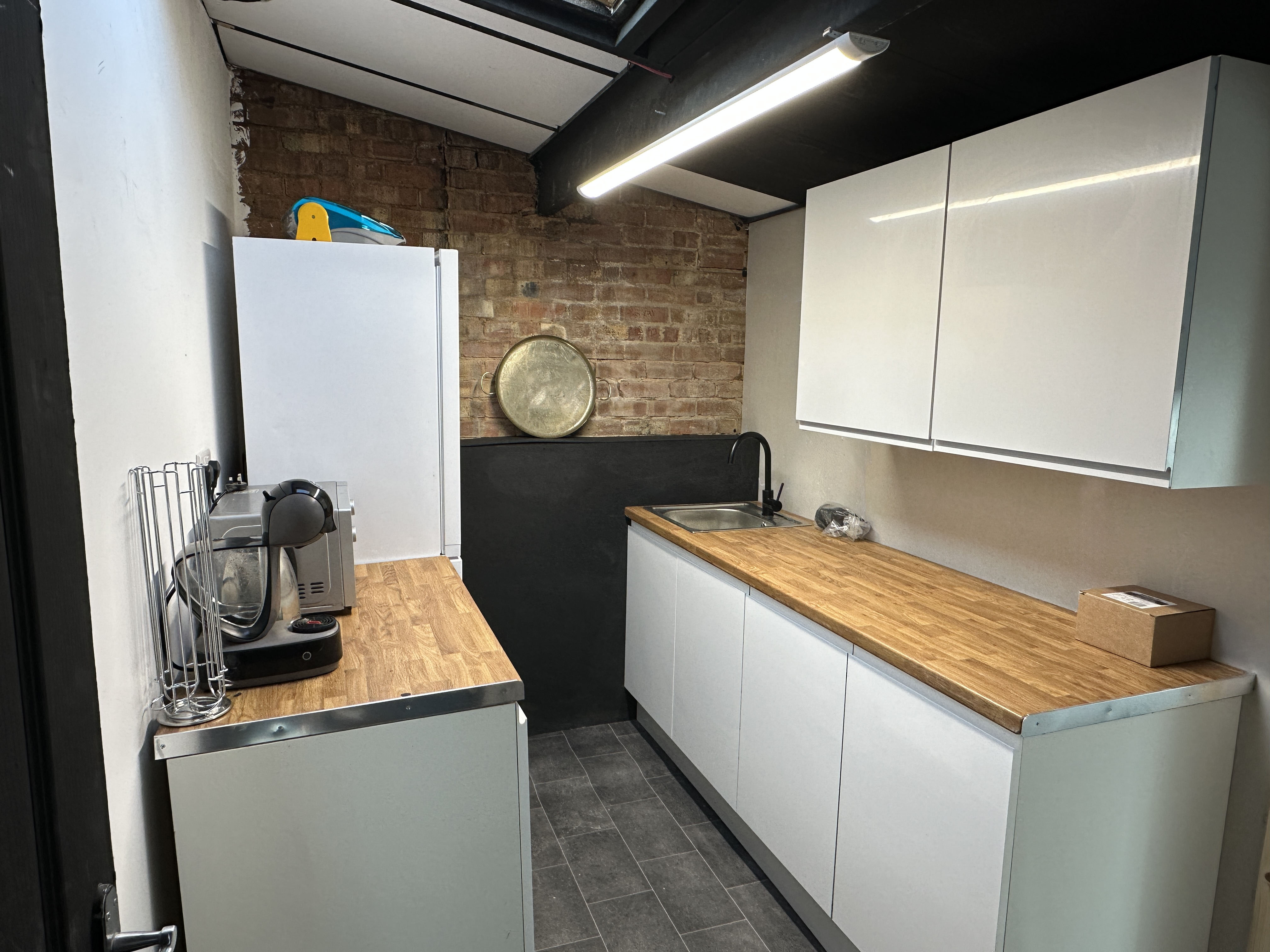 Modern narrow kitchen with white cabinets, wooden countertops, brick wall, and appliances including a coffee machine and microwave.