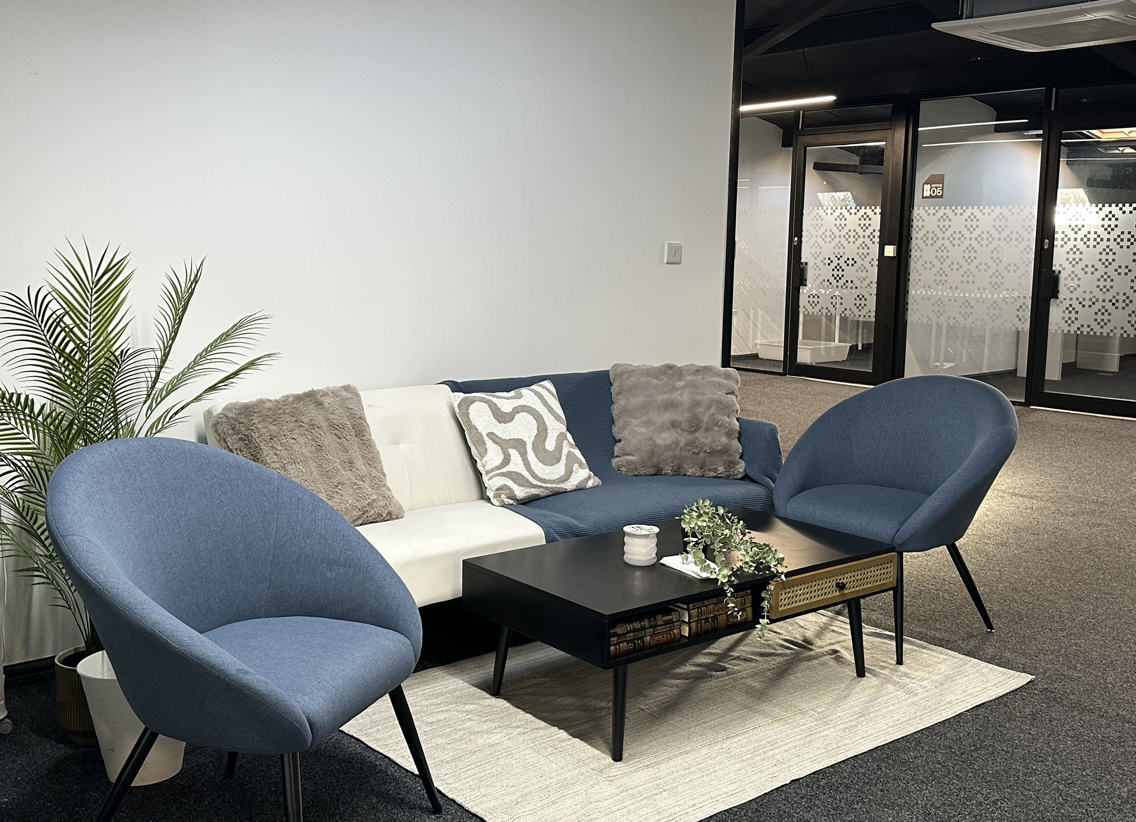 Modern office lounge with blue and white sofa, two blue armchairs, black coffee table with plants, and a potted plant against a white wall.