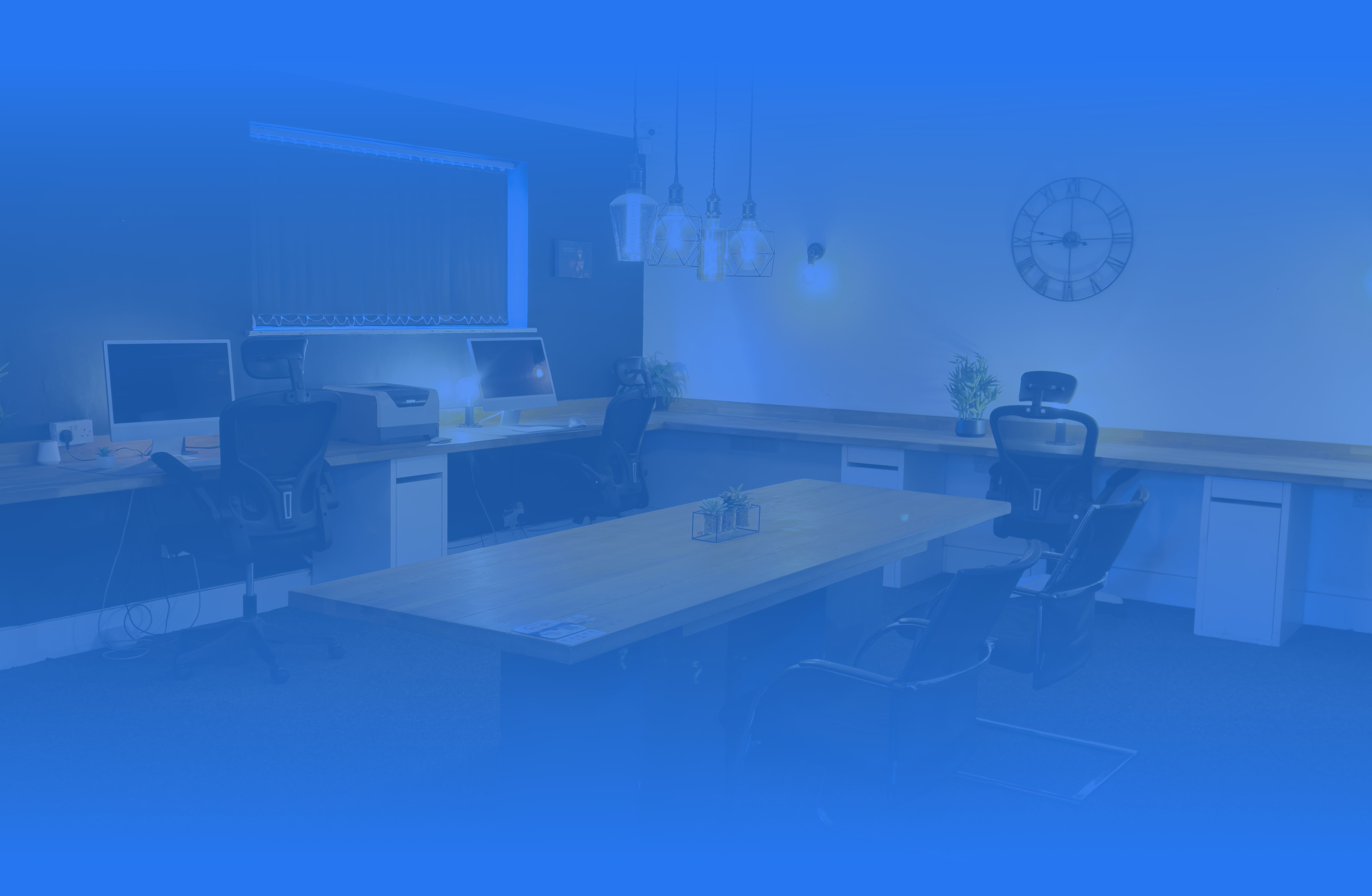 Modern office with wooden desks, ergonomic chairs, desktop computers, hanging pendant lights, and wall clock.