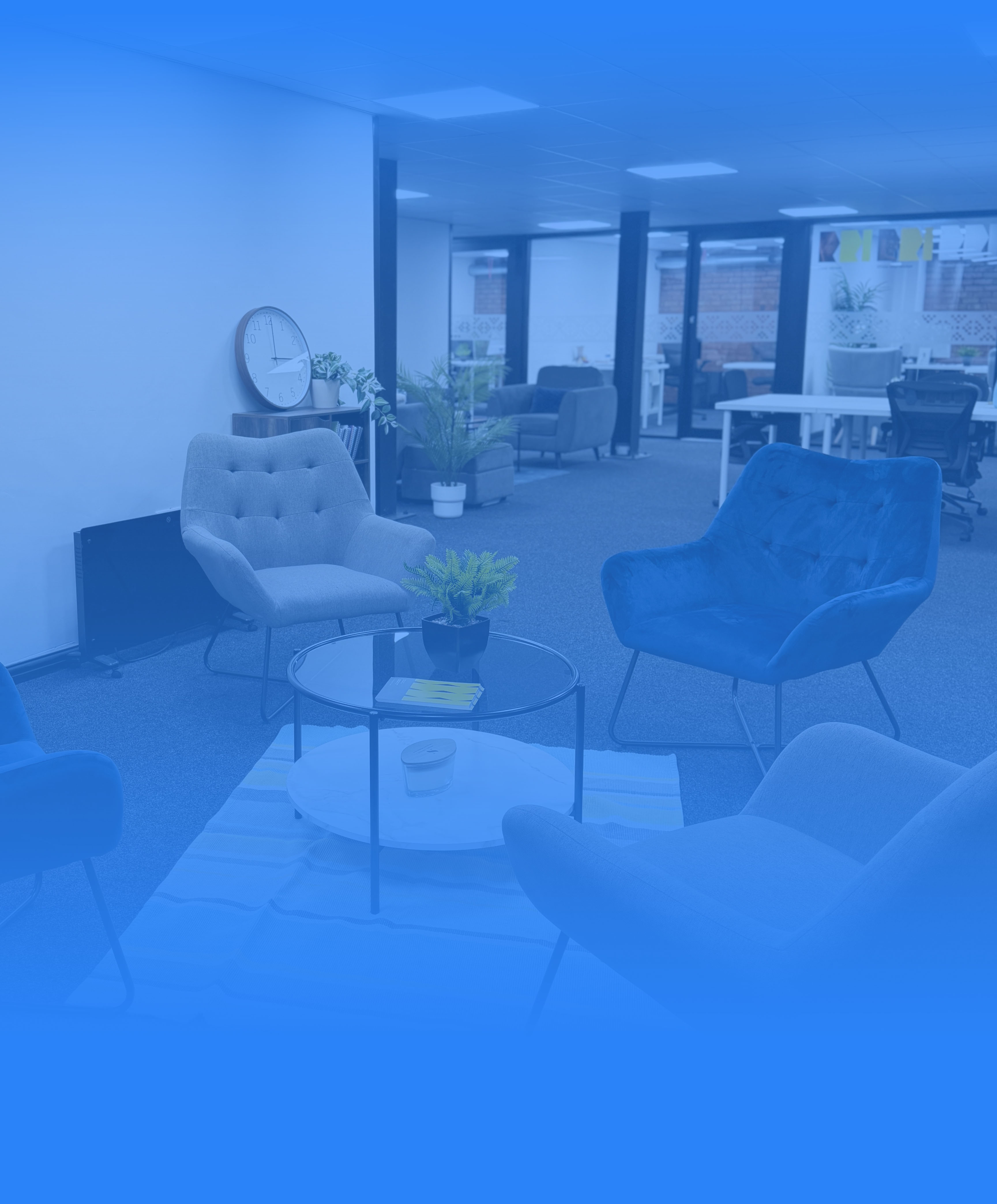 Modern office lounge area with four upholstered armchairs around a glass coffee table, potted plants, wall clock, and an open workspace in the background.