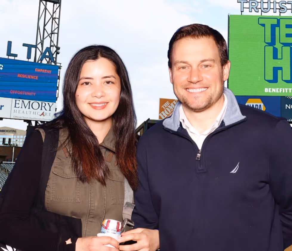 Smiling man and woman standing together outdoors in front of a stadium scoreboard.
