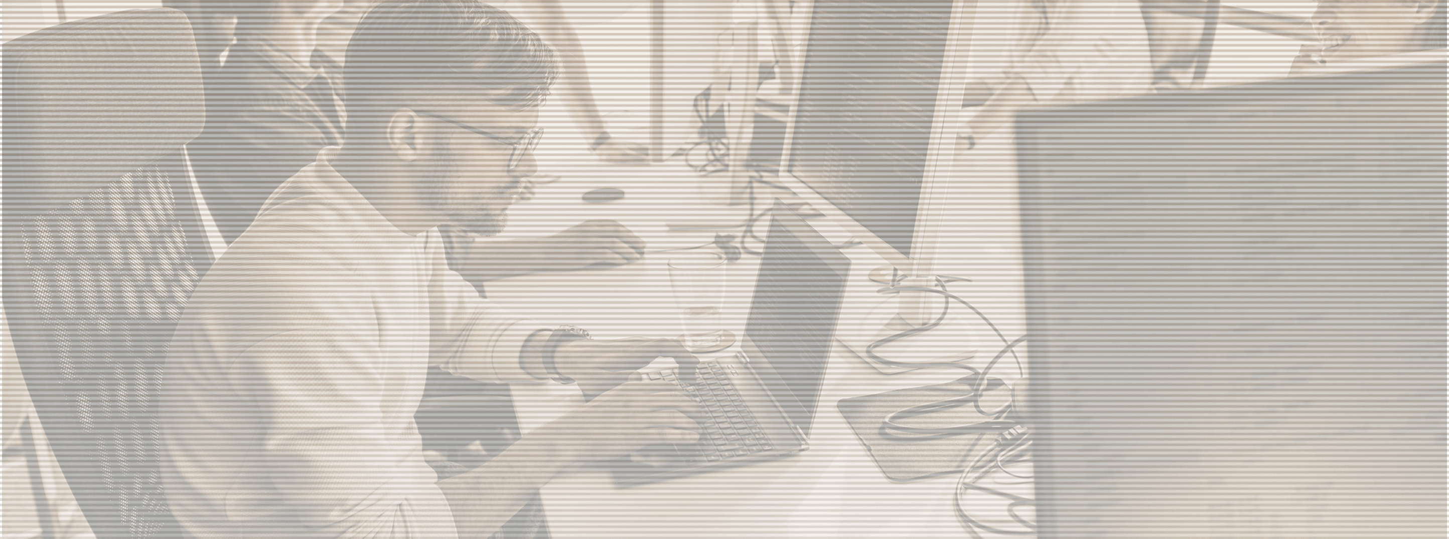 Man with glasses typing on a laptop at a desk in an office with multiple monitors and colleagues.