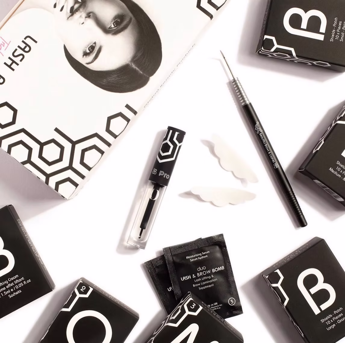 Flat lay of Duo Lash & Brow Bomb beauty products including serums, lifting cream sachets, applicator tool, and silicone shields on a white background.
