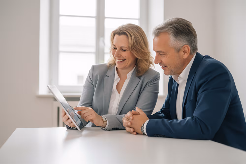 Zwei Geschäftsleute in Anzügen sitzen an einem Tisch, während eine Frau einem Mann etwas auf einem Tablet zeigt.