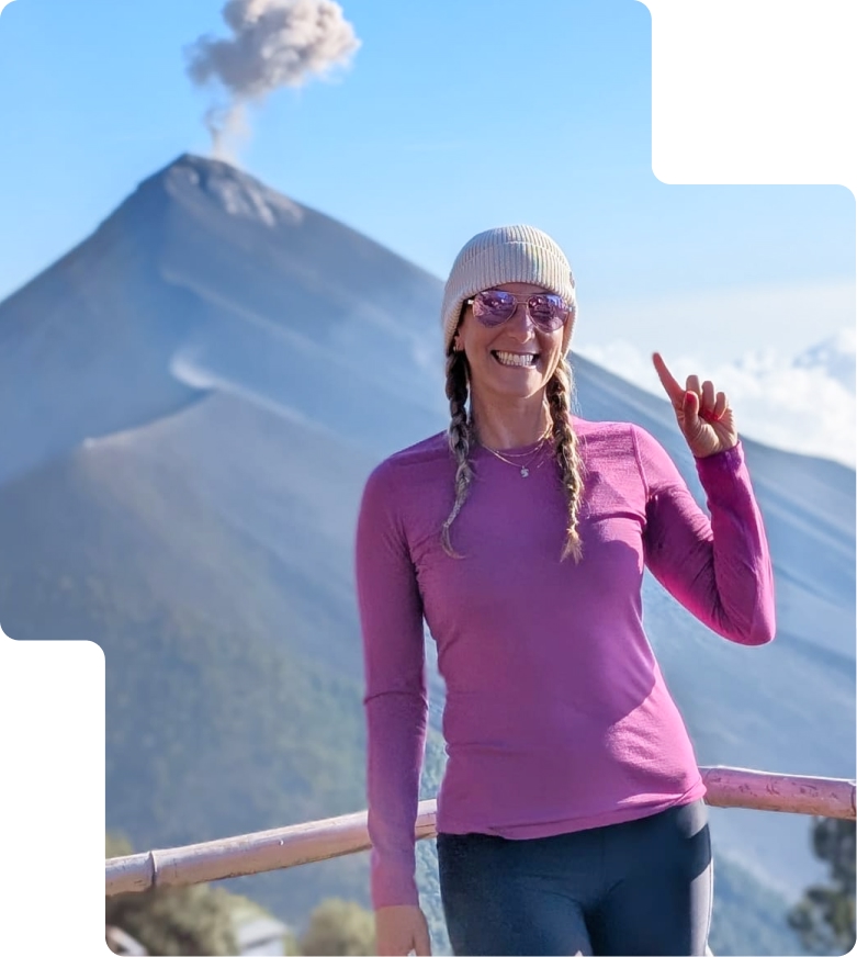 Kathryn in a pink sweater and beanie smiling and pointing at an erupting Mexican volcano in the background.
