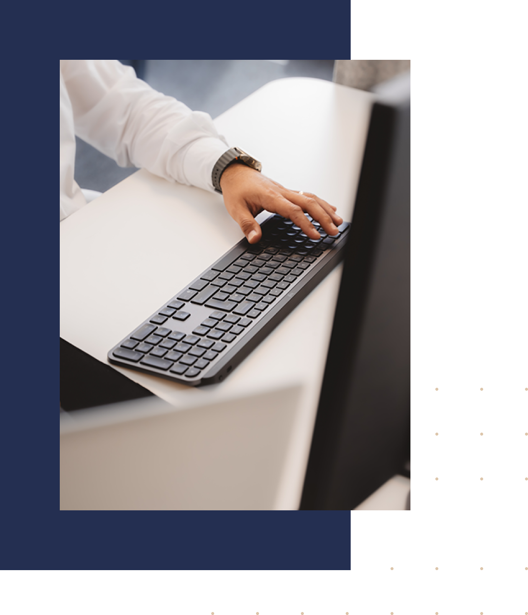 Main posée sur un clavier sans fil noir sur un bureau blanc devant un écran d'ordinateur.