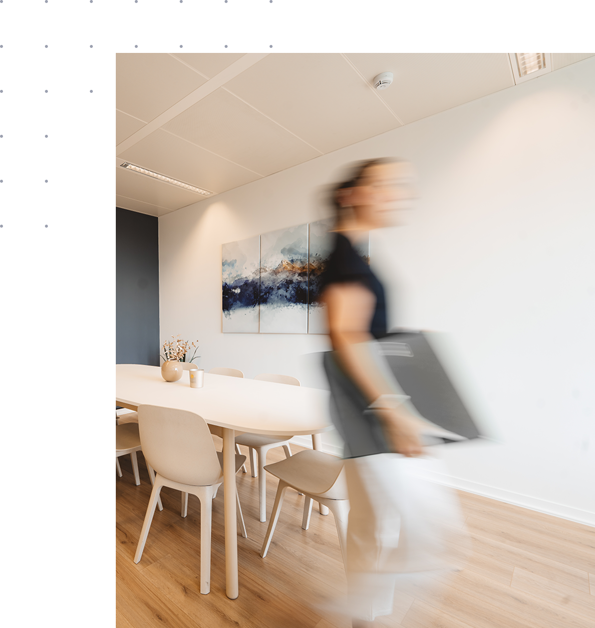 Salle de réunion moderne avec table blanche, chaises beiges, vase avec fleurs et tableau abstrait, une personne floue en mouvement.