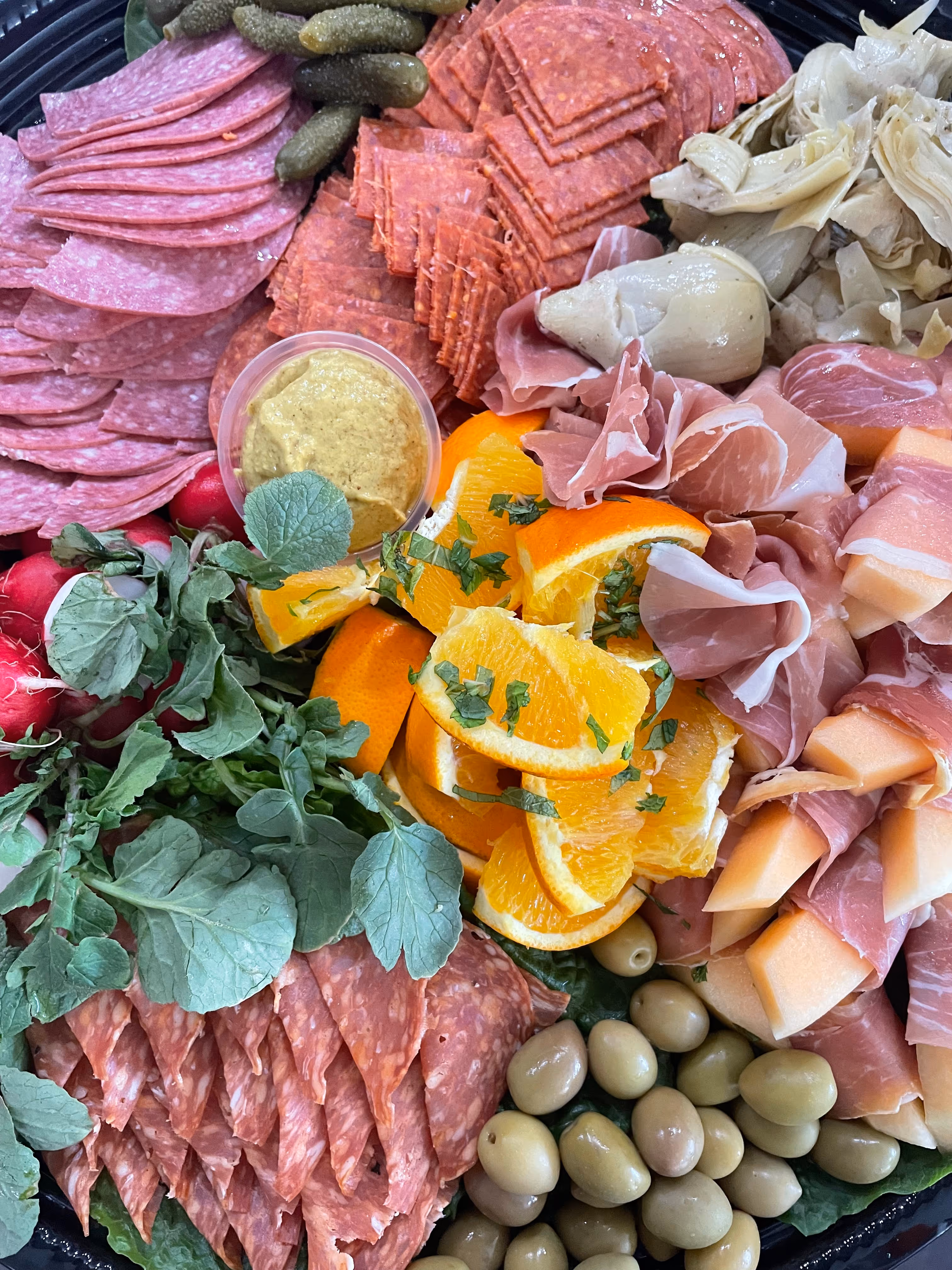 Charcuterie platter with sliced salami, prosciutto-wrapped cantaloupe, orange slices with herbs, green olives, pickles, radishes with greens, mustard, and artichokes.